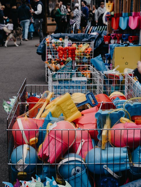 A metal wire basket filled with an assortment of plastic beach toys, including pink, blue, and yellow buckets, shovels, and sand molds, all made of hard, colorful plastic with smooth finishes. The toys are loosely piled inside the basket, some overlapping and slightly tilted, with a few spilling over the edges. The basket is placed outdoors on a paved surface, likely at a market or event, and is surrounded by other similar containers filled with various children's items. In the background, several people are visible, some browsing or talking, and additional market stalls displaying items such as gardening tools, shovels, and possibly other toys, with the scene lit by natural daylight. The image captures a moment typical of stallholders managing their merchandise, subtly illustrating the importance of independent waste handling and routine rubbish removal from market stall environments, consistent with Greenwich Market's setting and practices for stallholders to maintain clean, organized spaces while offering a variety of children's play items.
