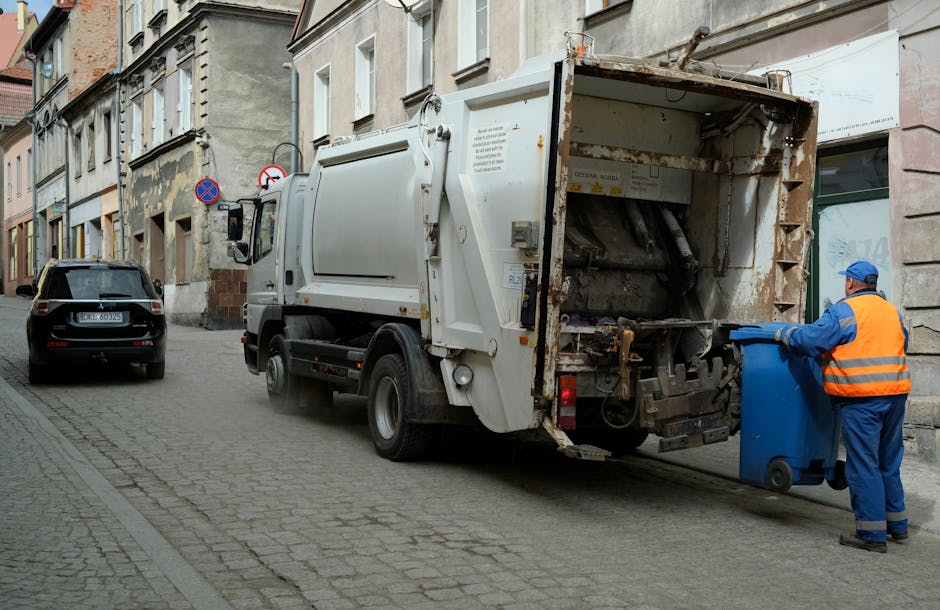 A street scene featuring a white waste collection truck with an open rear loading compartment, positioned on a cobblestone road adjacent to old multi-storey buildings with aged façades. The truck's rear section shows signs of rust and wear, with visible mechanical components and metal panels. The vehicle is operated by a worker dressed in blue and orange high-visibility clothing, standing to the right, actively pushing a blue wheeled bin towards the truck’s opening. In the background, a black car is parked parallel to the curb, and there are street signs attached to the building façade indicating parking restrictions. The scene occurs during daylight under an overcast sky, providing neutral, diffuse lighting, typical for waste collection operations in urban areas. This image reflects a routine rubbish removal process, possibly part of a private or independent waste management service, supporting the context of rubbish and waste disposal for local businesses or residents.
