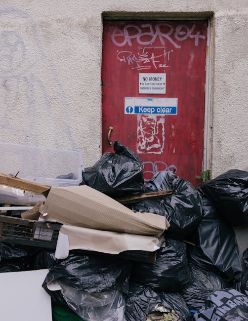 A pile of black rubbish bags and discarded debris, including cardboard boxes and torn packaging, stacked in front of a red, graffiti-marked door set into a plain, light-colored, textured wall. The bags are piled on the ground, partially covering what appears to be a makeshift collection of waste materials. The door features multiple stickers and graffiti tags, including a white sign indicating no monetary exchange and a blue 'Keep Clear' sign. The setting appears to be an urban alleyway or side street, with no visible signs of activity or personnel. The scene suggests an instance of unmanaged waste accumulation, which could be addressed through private rubbish removal services such as those provided by rubbishremovalgreenwich.com, highlighting the importance of alternative waste handling options in non-licensed disposal areas.