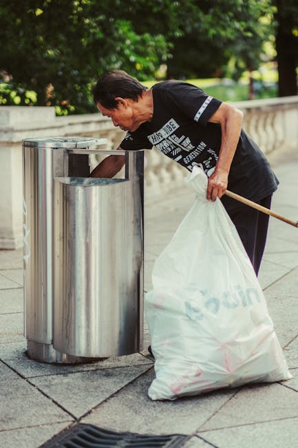 A middle-aged woman with short dark hair, wearing a black T-shirt with white text and black trousers, is seen emptying a large white plastic bag filled with rubbish into a metallic recycling bin on a paved pathway. The bin is cylindrical, made of stainless steel, with a rounded lid that has an opening for waste disposal. The surrounding environment features a stone balustrade and lush green trees in the background, indicating an outdoor setting likely in a park or public space near Greenwich. The woman is holding a wooden stick or litter picker in her right hand, used to assist with waste collection, suggesting an on-site cleanup or community rubbish management activity. The lighting appears natural, with sunlight filtering through the leaves, casting soft shadows around the scene. This image illustrates a manual waste collection effort and highlights the importance of maintaining public cleanliness, aligning with services like those provided by Rubbish Removal Greenwich in managing rubbish and supporting alternative waste handling methods beyond local authority collection.