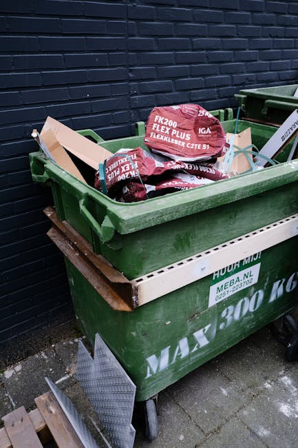 A large green wheeled rubbish skip filled with red and black plastic bags containing building or garden waste, positioned outdoors against a dark blue brick wall. The skip rests on a paved surface, with a white plastic panel attached to its side displaying contact details and waste capacity information. Nearby, a smaller green bin is partially visible, along with some construction or garden tools resting against the front of the skip. The scene is lit with natural light, highlighting the textures of the plastic bags, metal rim, and brick wall, indicating an environment suitable for private waste collection or on-site rubbish removal services such as those offered by Rubbish Removal Greenwich, aligned with alternative waste handling methods for residential or commercial waste clearance.