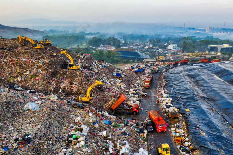 A large pile of mixed rubbish is accumulated on a paved urban street near a parking area, positioned behind a metal railing with a small tree on the left side. The rubbish includes various materials such as flattened cardboard boxes, plastic bags filled with waste, paper, and household items. Some cardboard boxes are partially collapsed and several black and red refuse bags are visibly tied and stacked among the clutter. A prominent grey metal skip, labeled for mixed paper and cardboard disposal, holds more loose waste and is surrounded by overflowing trash, with some waste spilling onto the ground. To the left of the skip, a small silver hatchback car is parked parallel to the curb, next to a green and white waste collection point or recycling station visible in the background. Behind the rubbish, there is a building with a blue scaffolding structure, and storefronts with partially visible signage and yellow and green accents, suggesting a commercial or retail area. The scene conveys an example of improper waste disposal or a temporary accumulation outside a waste management service like Rubbish Removal Greenwich, situated in a busy urban environment.
