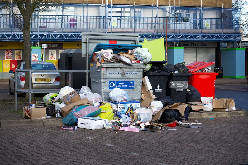A large collection of waste and rubbish is piled on a city pavement near a parking area, including overflowing black and red plastic rubbish bins, with some lids open, revealing mixed waste materials such as crumpled paper, cardboard boxes, and plastic bags. Cardboard boxes are scattered around the base of the bins, some flattened and others intact, while various paper and packaging waste are strewn across the ground. In the background, a grey van is parked behind a metal safety barrier, with a multi-storey building featuring blue scaffolding and signs for local businesses visible behind it. The environment appears to be an urban setting with minimal greenery, as a leafless tree stands adjacent to the waste pile, indicating a colder season. The scene illustrates a typical instance of excess waste awaiting collection, which companies like Rubbish Removal Greenwich might handle through private on-site clearance or alternative waste disposal services, especially in areas where street refuse collection is limited or delayed.
