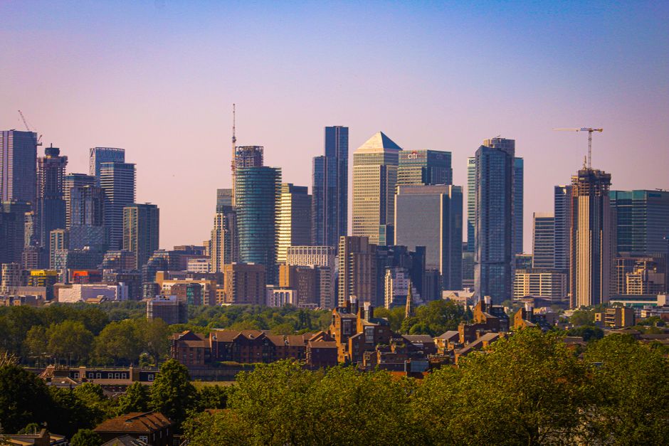 A city skyline featuring a variety of modern high-rise buildings with glass facades reflecting sunlight, situated behind a foreground of dense green trees and residential houses with pitched roofs. The skyscrapers include rectangular, tapered, and triangular shapes, some under construction with visible cranes. The scene captures a clear, blue sky with soft light, suggesting daytime, and overall reflects an urban environment with a mix of commercial and residential architecture, related to the context of rubbish removal services by companies specializing in private waste clearance or on-site rubbish collection near Greenwich riverside estates.